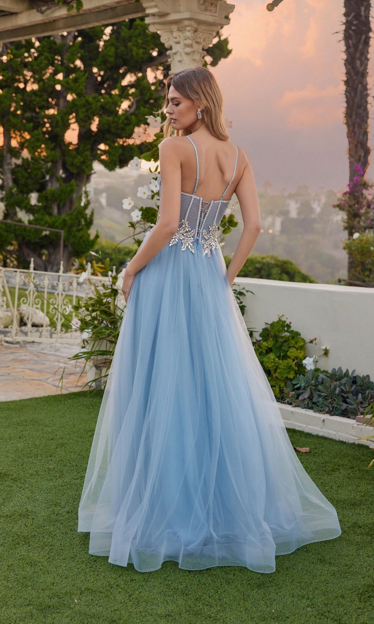 Wearing the Juliet JT2638G, a woman stands outdoors on green grass at sunset. Her light blue, floor-length A-line prom dress features a tulle skirt and floral embellishments on the back near a garden railing.